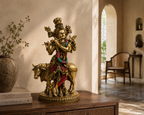 Krishna idol with cow displayed on a wooden console in a sunlit interior beside books and a vase