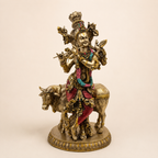 Krishna idol playing a flute beside a cow, with gold-tone finish, red clothing and a pink and turquoise garland, front view
