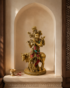 Krishna idol playing a flute beside a cow in an arched prayer niche with a diya and flower petals