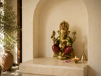 Seated Ganesha idol displayed in a sunlit niche with a diya, bell and flower petals