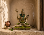 Meditating Shiva idol displayed in a sunlit stone shrine with a diya, copper-tone vessel and incense